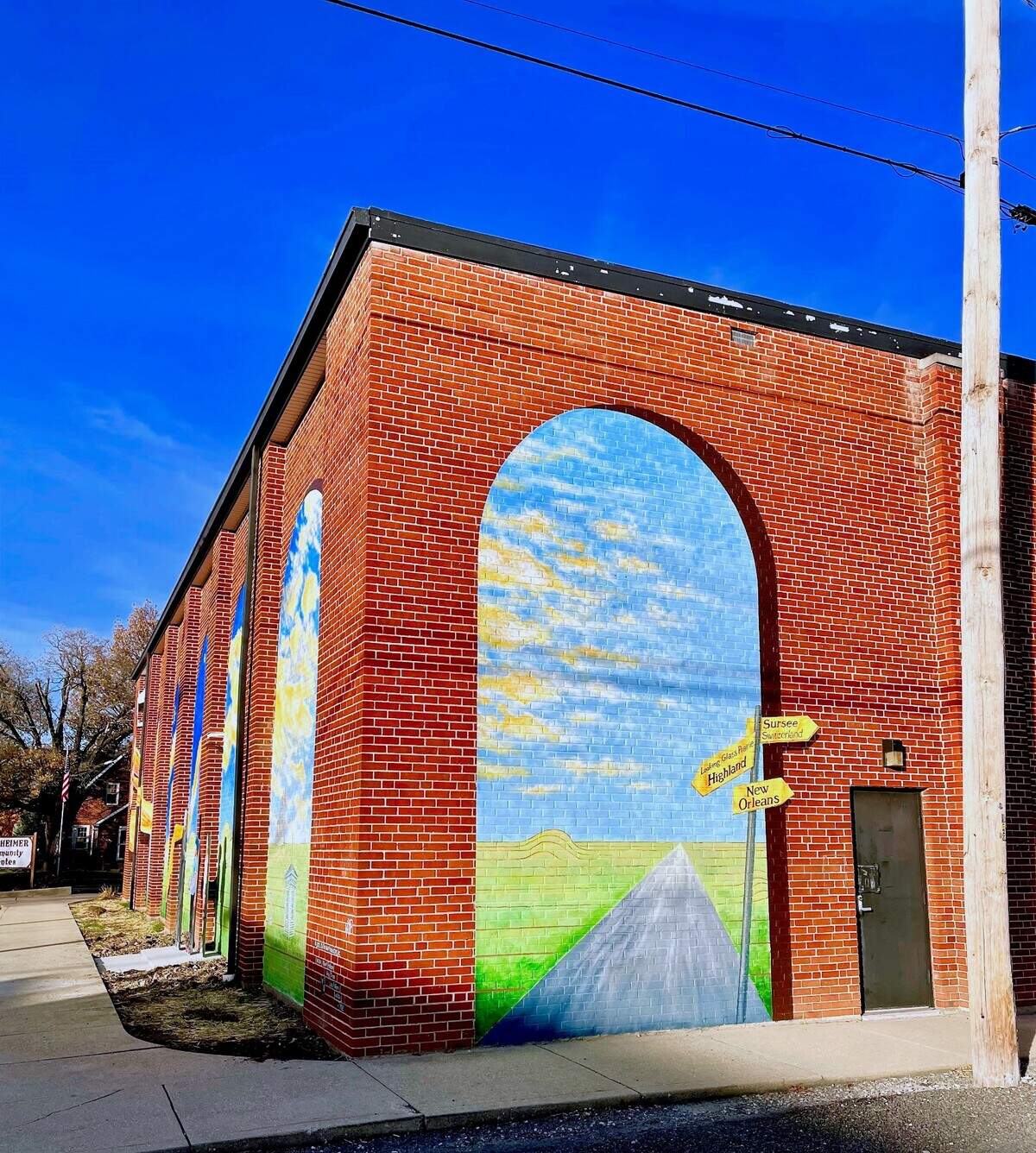 Made possible in part by a $10,000 matching grant from the National Endowment for the Arts, a mural was designed and painted on the side of the Weinheimer Community Center at the corner of Main and Pine Streets in Highland, by Robert and Liza Fishbone of On the Wall Productions from St. Louis. They were assisted onsite by Andy Cross (set painter for The St. Louis Muny Opera), and Lisa Roth. Deborah “Moe” Moellering painted the three cut out figures installed next to the mural.