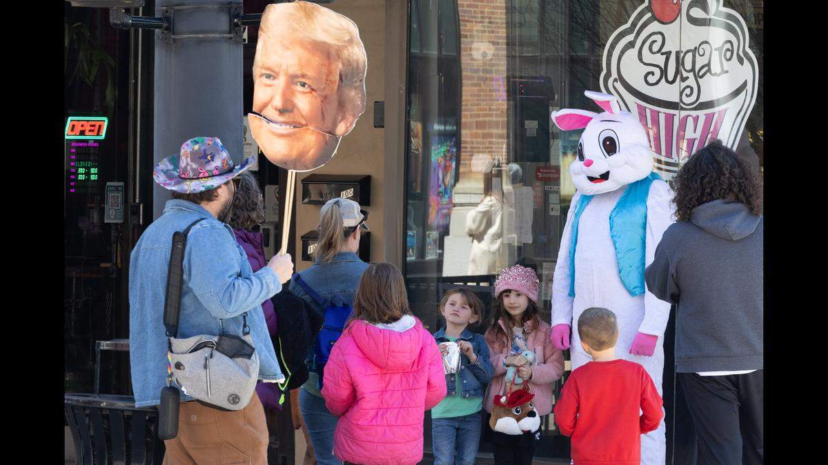 A No Kings protester holding a Donald Trump sign with a movable mouth stops in front of Sugar High Bakery & Cafe in downtown Belleville as children chat with the Easter bunny.
