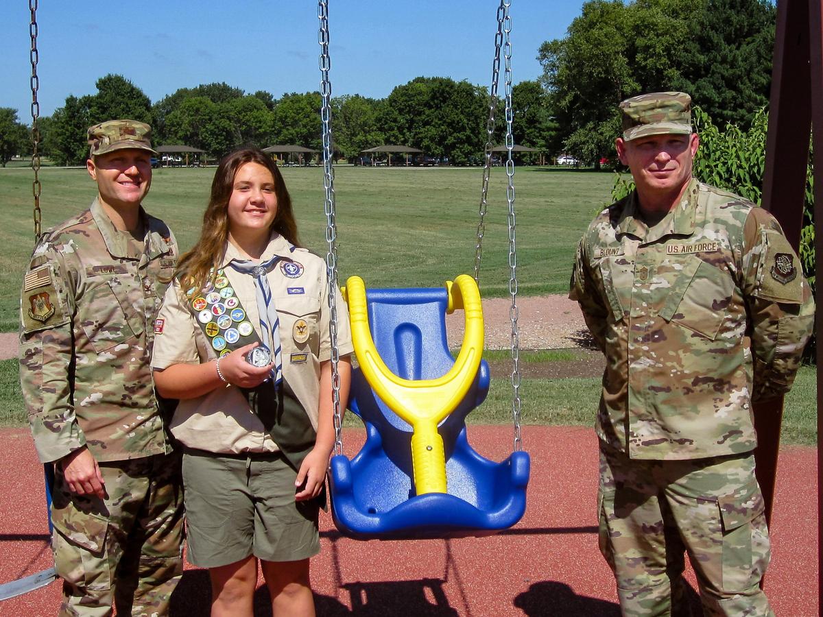 Elise Toth, Boy Scouts of America Troop 144 member, is presented a coin by Col. Rob Lowe, 375th Air Mobility Wing vice commander and Chief Master Sgt. Thomas Blount, 375th AMW command chief, at Warrior Park on Scott Air Force Base. In support of her Citizenship in the Community Badge, Toth inquired with a community leader as to why none of the installation playground swings were compliant with the Americans with Disabilities Act. After jumping through several hurdles to secure funding, Richard Cole, 375th Civil Engineer Squadron installation management chief and Mark McCoy, 375th CES pollution prevention and cultural resource manager, were able to purchase the swing by classifying it as an outreach project. Thanks to Toth, the new swing was installed Aug. 10 and is now available to the installation.
