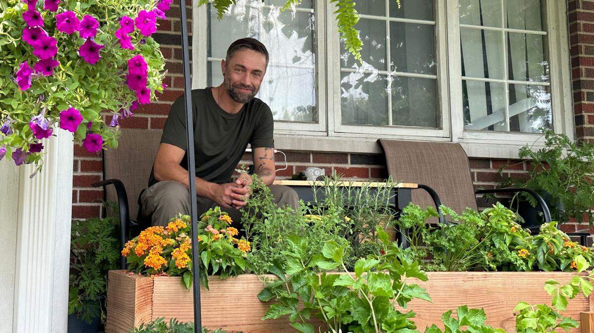 Ryan McCann stares and smiles at the camera while sitting on a chair in front of flowers and plants.