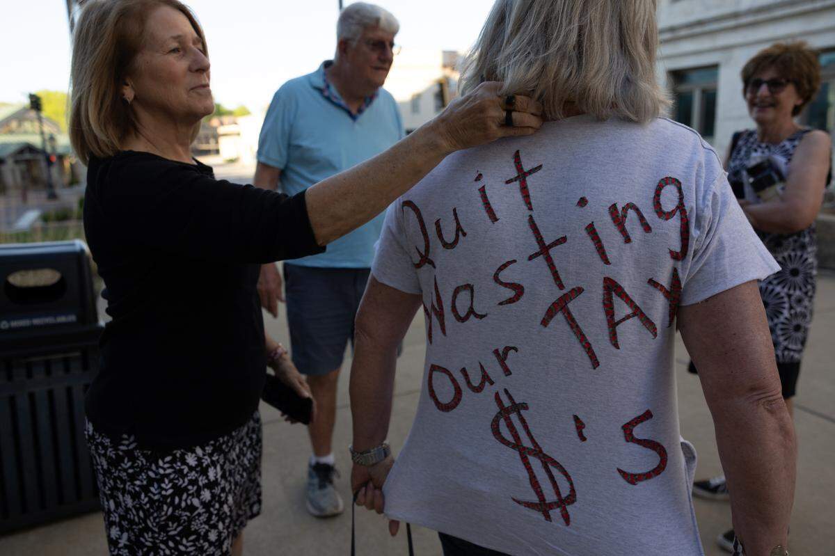 Sue Betts (L), 71, helps friend and fellow Edwardsville resident Sheri Schuler-Faust, 69, tuck her scarf into her collar so her message of “quit wasting our tax $’s” can be more easily read after a Madison County Board meeting on April 17, 2024.