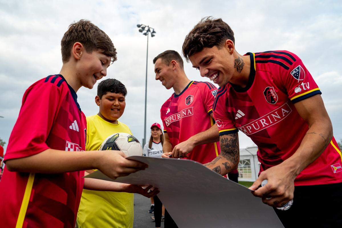 St. Louis City SC midfielder Célio Pompeu (12) signs an autograph on a poster held by Noah White, 12, of Collinsville, on Thursday, Oct. 26, 2023, during the dedication of a St. Louis City SC mini pitch at Granby Park in Fairmont City. Israel Aguirre, 10, of Collinsville, watches while standing next to St. Louis City SC defender Kyle Hiebert (22).