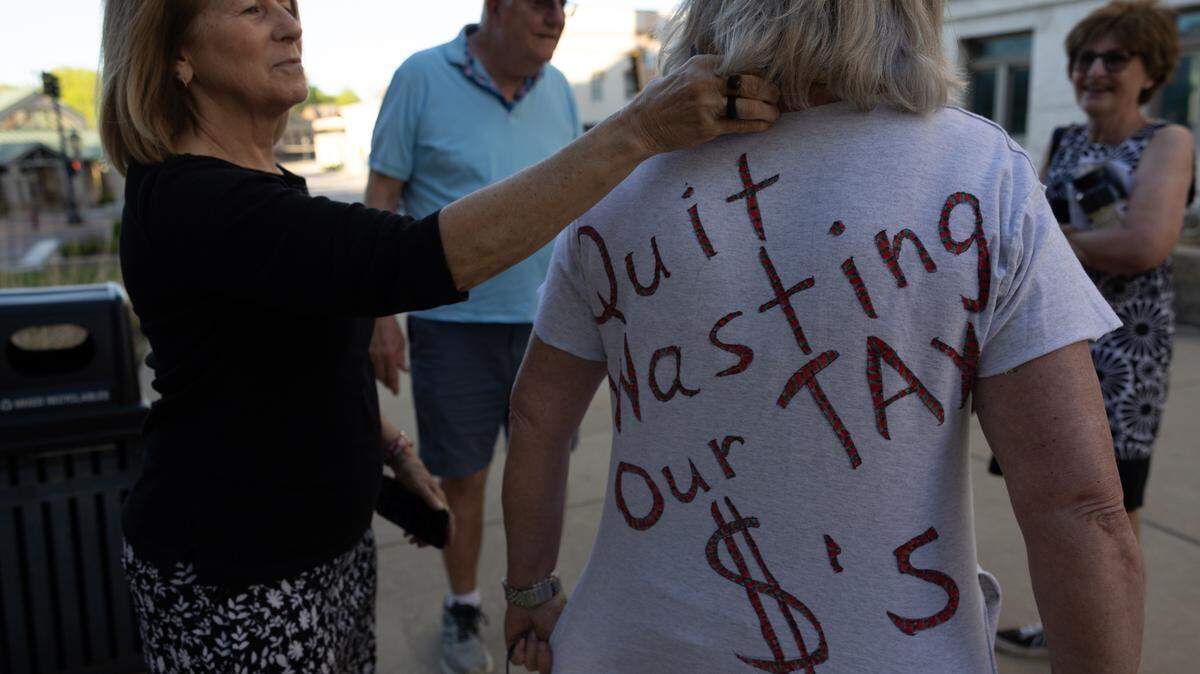 Sue Betts (L), 71, helps friend and fellow Edwardsville resident Sheri Schuler-Faust, 69, tuck her scarf into her collar so her message of “quit wasting our tax $’s” can be more easily read after a Madison County Board meeting on April 17, 2024.