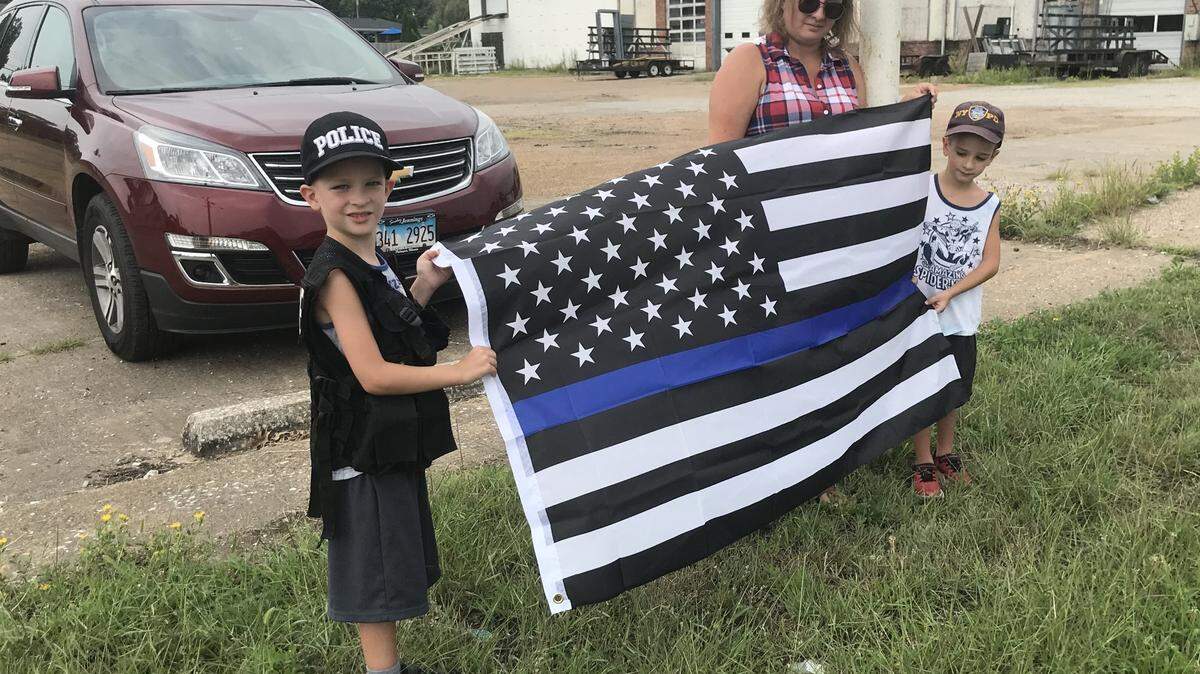 Julia Campbell and her 6-year-old twin sons, Colton, left, and Cooper, hold a “thin blue line” American flag along the route of a procession honoring fallen Brooklyn police officer Brian Pierce in Wood River on Friday.