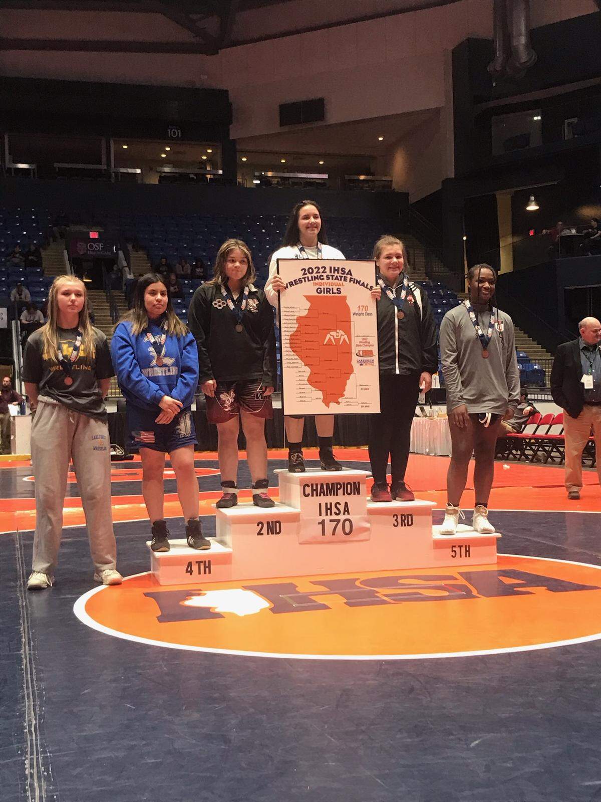 Highland girls wrestler August Rottmann stands on the medal stand (right of top) after placing third in the 170 division of the inaugural IHSA girls individual state tournament on Saturday, Feb. 26 at Grossinger Motors Arena in Bloomington.