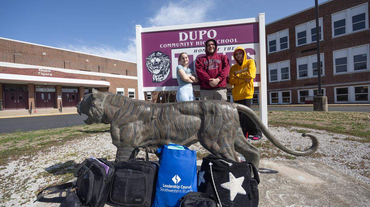 From left to right, Dupo High School junior Shaylee Messersmith, junior Dylan Cannon and sophomore Jasper Anderson pose in front of the school sign with the bags the students carry in lieu of a backpack. Messersmith explained the policy restricts the size of the bags students can carry from class to class.