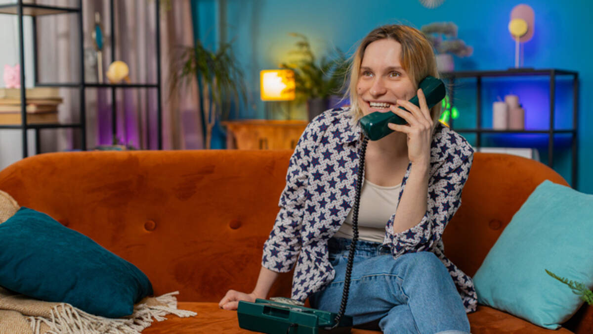 Digital detox is shifting toward everyday habits that help people spend less time on their phones without giving them up completely. Smiling young woman making wired telephone call conversation with friends sitting on couch at home in room. Happy excited pretty girl enjoying old-fashioned retro phone from 90s talking in living room