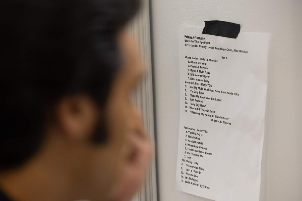 An Elvis tribute actor puts on makeup while a setlist for other Elvises is posted in the background.
