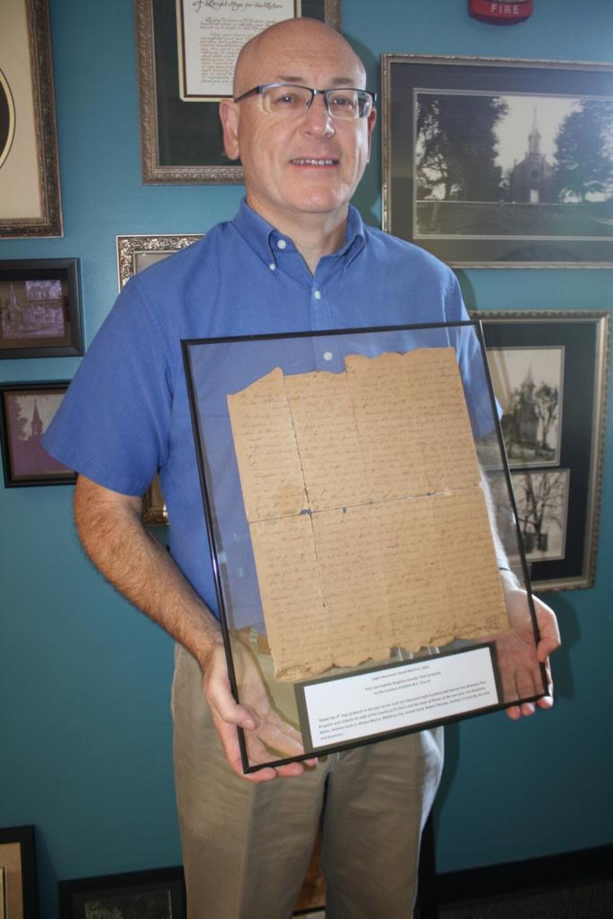 Shiloh United Methodist Church Pastor Ken Hutchens stands in front of the historic wall located between the 1875 church chapel and the 2001 addition highlights historical documents, photographs and artifacts of the church’s long 210-year history. Hutchens holds a legal document dated March 4, 1822 for the transfer of property of Paul and Isabella Kingston to the trustees of Shiloh M.E. Church.