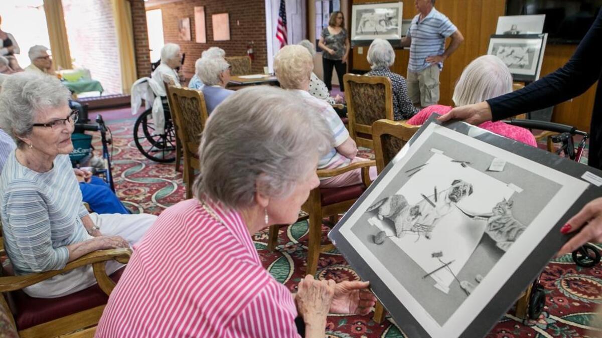Shirley Sargeant and Charlotte Yost take a close look at the work of graphite pencil artist Robin Lauersdorf. Lauersdorf, in the background, speaks to residents at the National Shrine of Our Lady of the Snows retirement living community. Lauersdorf speaks about his art career and the long process it takes to complete his current style of whimsical pencil drawings. Lauersdorf was one of seven artists participating in the Art on the Square Artist in Residence Program.