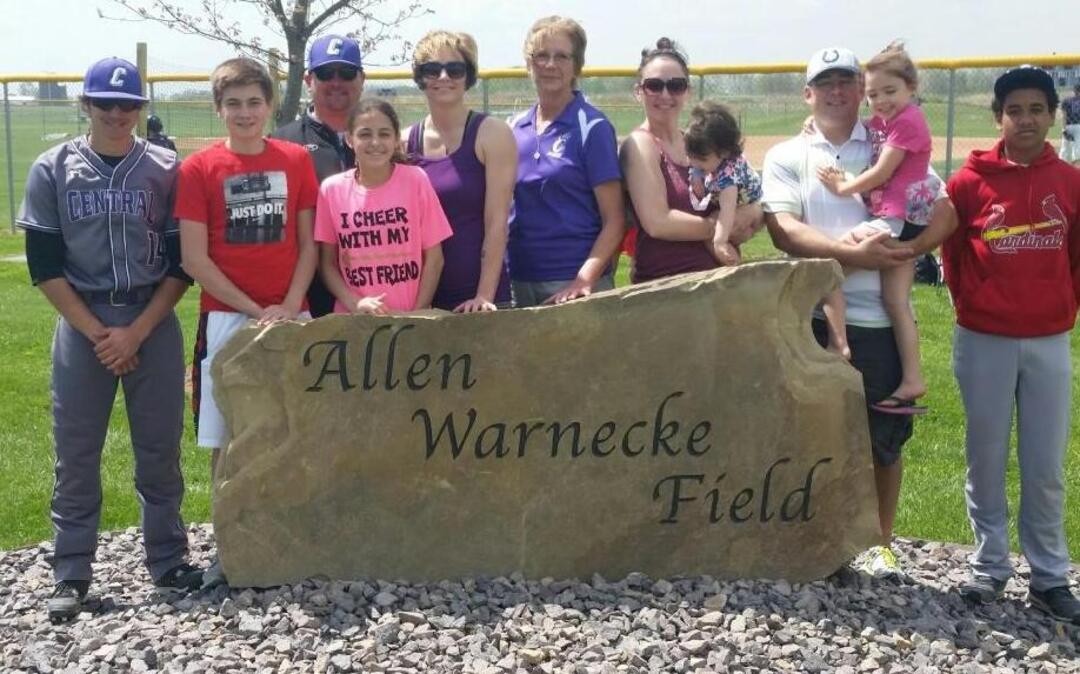 Pictured wih the new Allen Warnecke Field sign that was dedicated Saturday are, from left, Jayce Timmermann, Jeremy Timmermann, Jason Timmermann, Jadyn Schulte, Sarah Timmermann, Diane Warnecke, Jenny Santel (holding Norah Santel), Greg Santel (holding Charlie Santel) and Nicholas Warnecke.